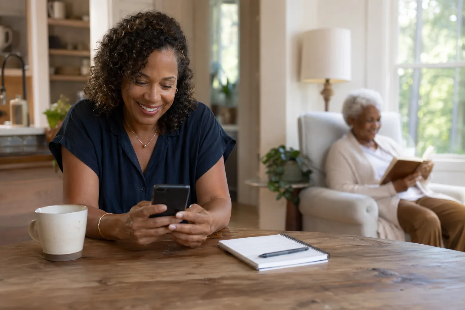 A woman smiling at her phone while her elderly mother reads in the background